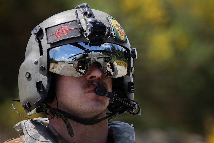 A close-up view of a man wearing an aviation helmet with a reflection in the visor of a military helicopter parked on the ground nearby.