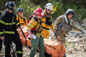 A man wearing a camouflage military uniform and aviation helmet and four other men in firefighter uniforms carry equipment outside.