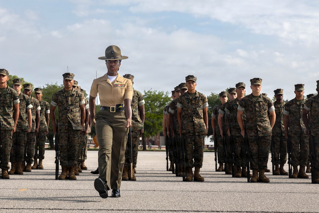 A Marine leads dozens of recruits carrying rifles while standing in formation during the day.