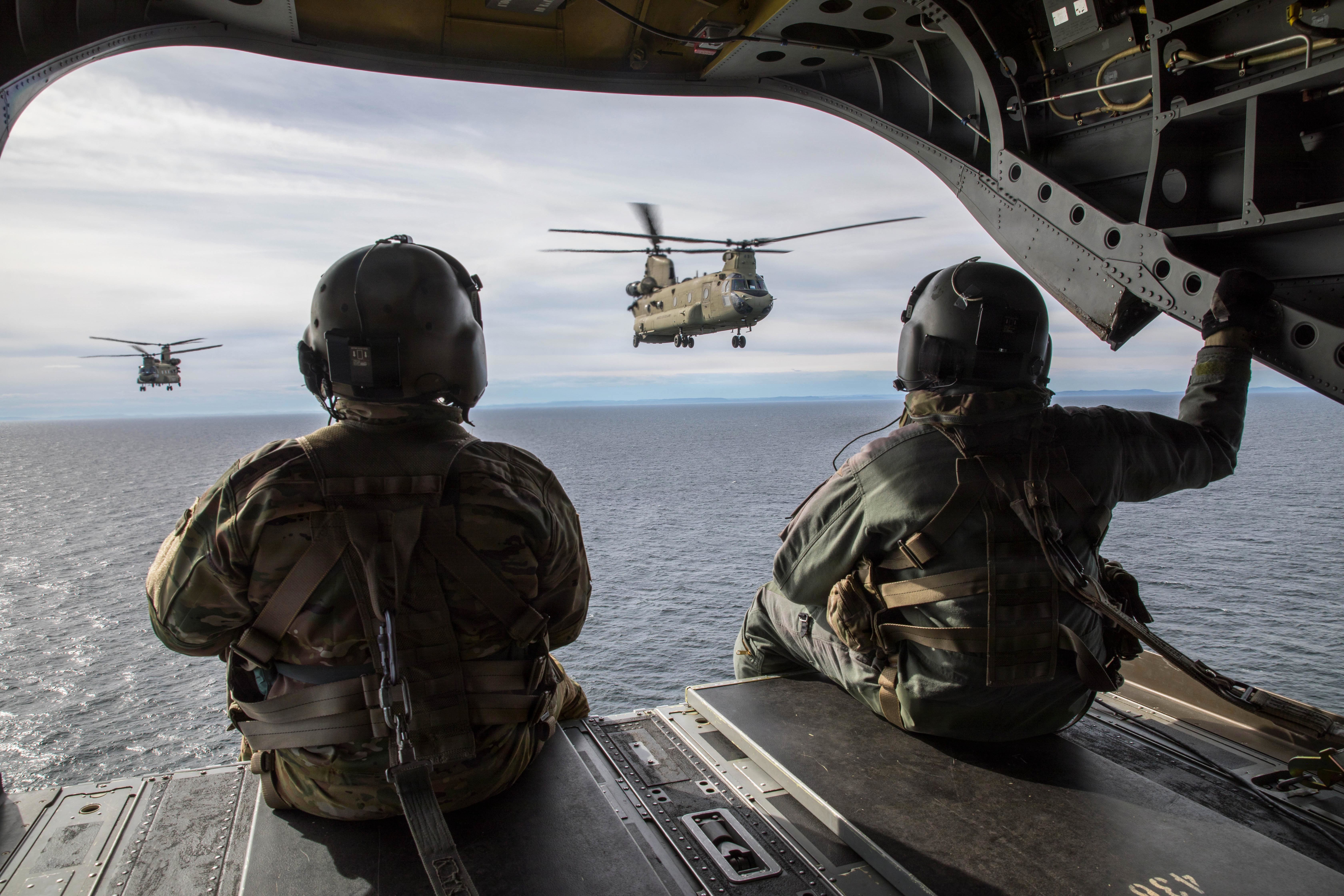 Two service members look out the open back of an in-flight aircraft toward two nearby helicopters flying over water.