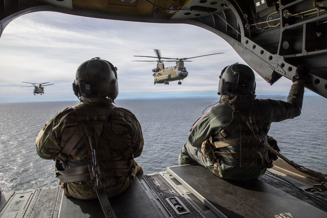 Two soldiers in tactical gear sitting in the open doorway of an aircraft as it flies over a body of water watch while two aircraft fly in the distance.