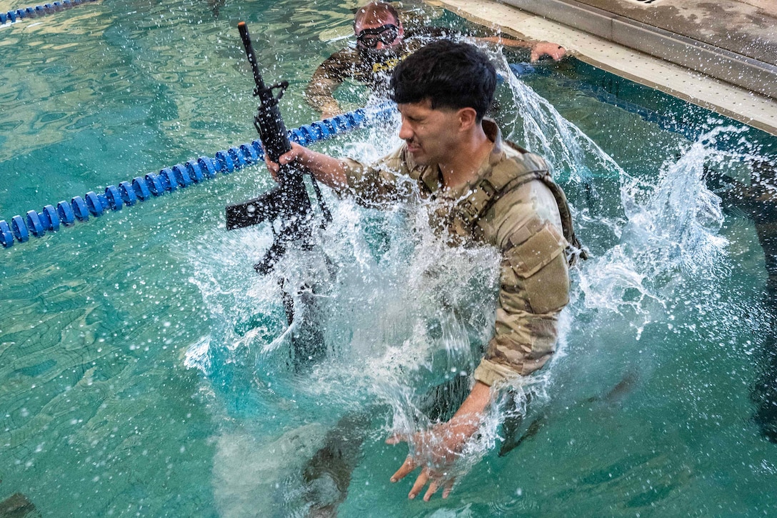 A soldier in tactical gear jumps into a pool with a weapon, causing a large splash as another soldier watches.