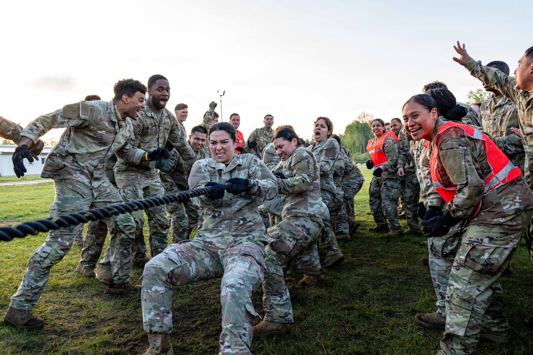Soldiers pull a rope as fellow soldiers cheer for them on a field during the day.