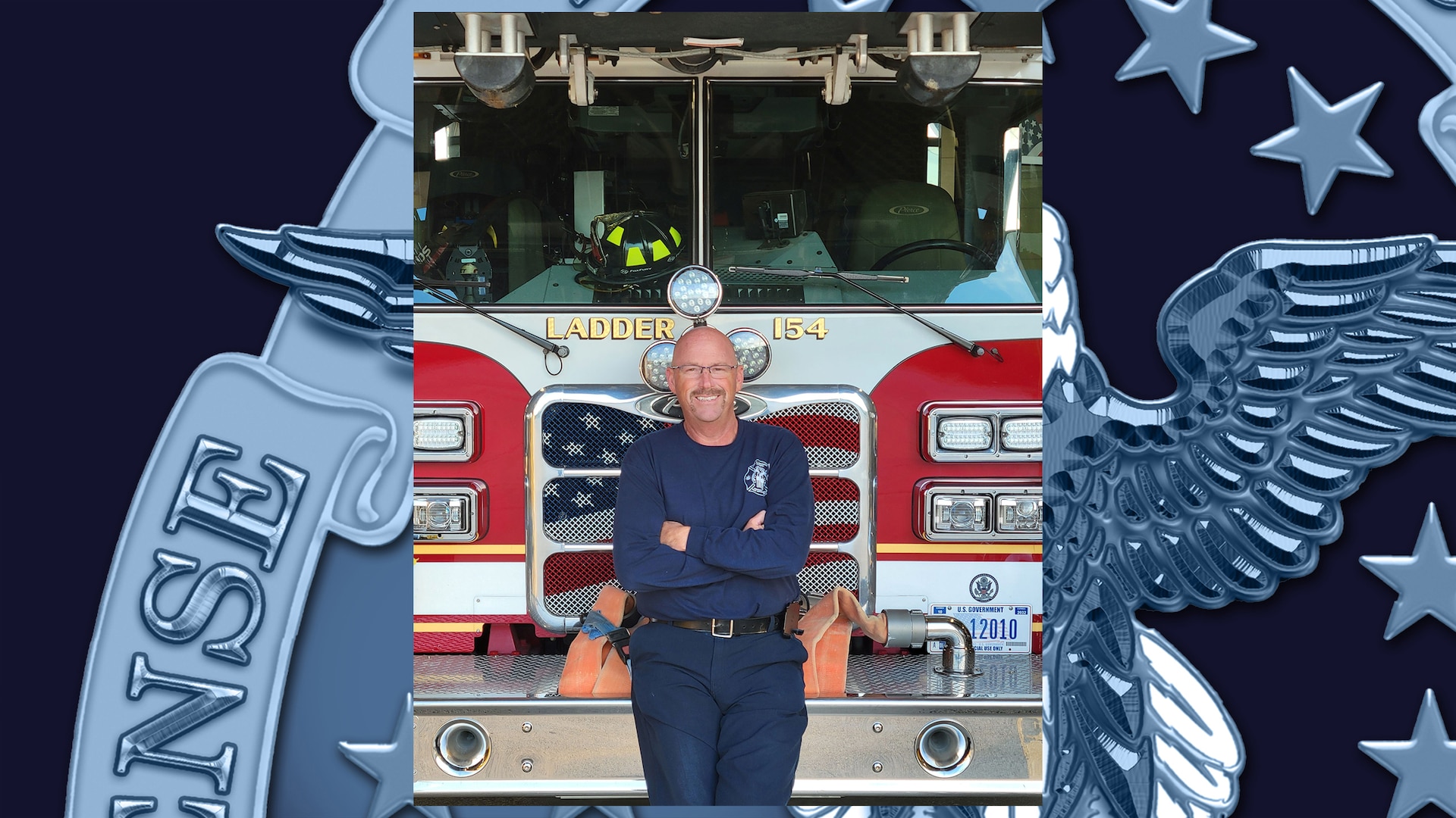 Man in blue pants and shirt, mustache, eye glasses and bald head smiles and poses in front of fire truck with arms crossed.