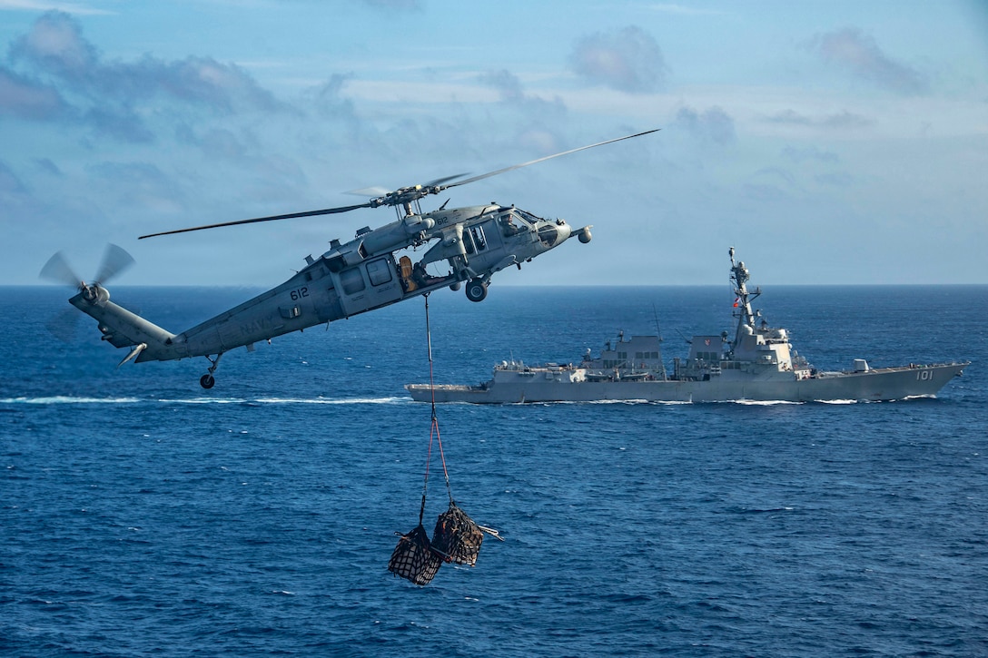 A helicopter carrying two loads of unidentified items in nets hanging from it flies over water, with a ship, clouds and the horizon in the background.