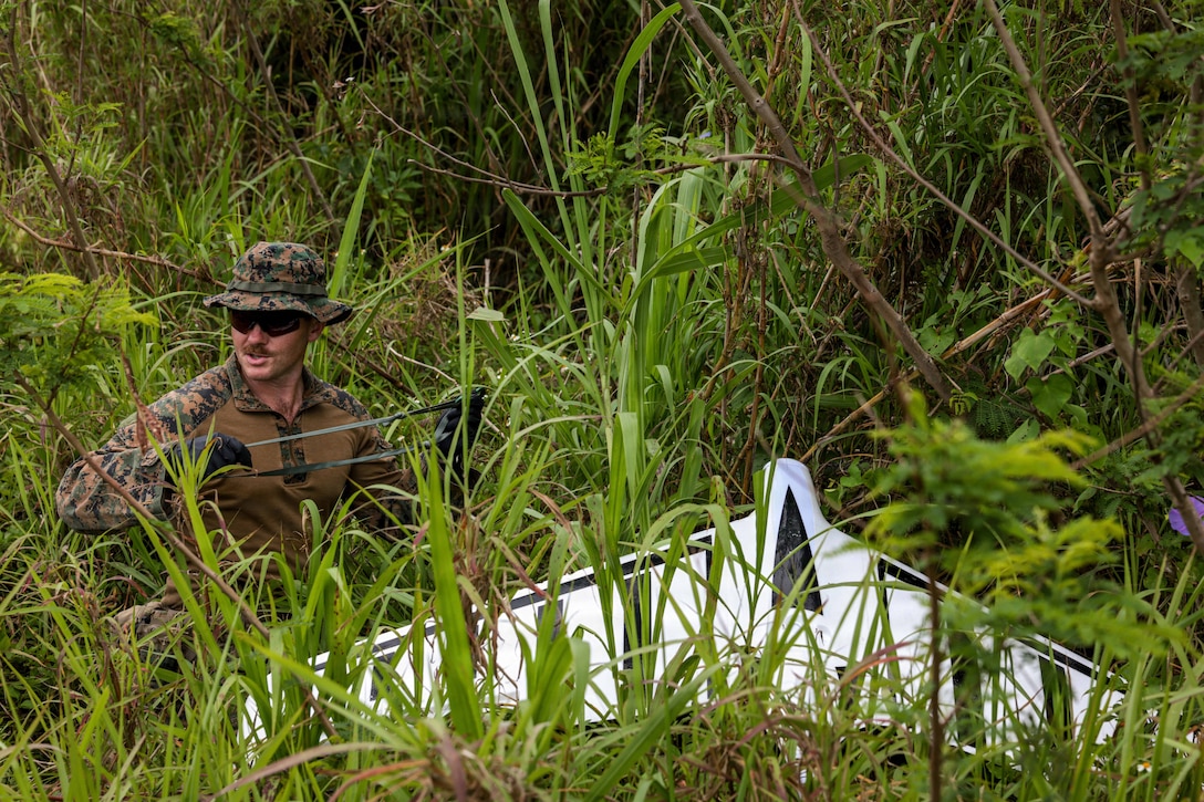 A soldier and a medium-size white drone with black markings are shown in tall grass and weeds.