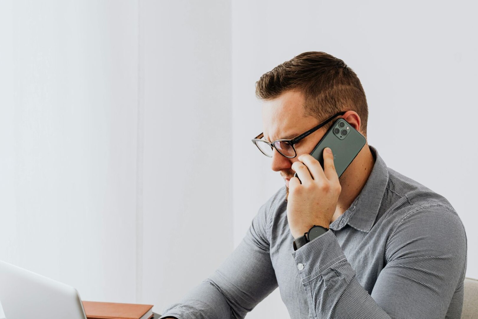Man in gray shirt talks on the phone while looking at a laptop.
