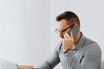 Man in gray shirt talks on the phone while looking at a laptop.
