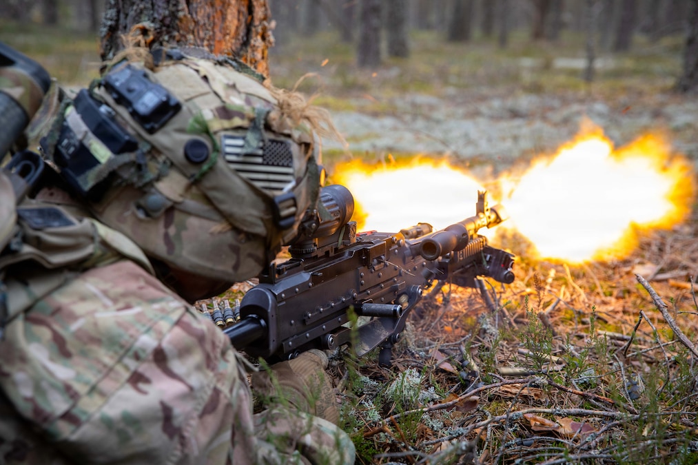 A soldier fires an assault rifle while low to the ground, with grass and tree stalks in the background.