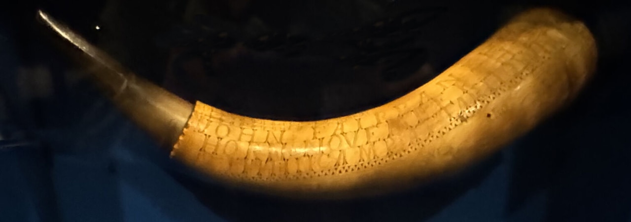 A golden color powder horn with engravings is on display.