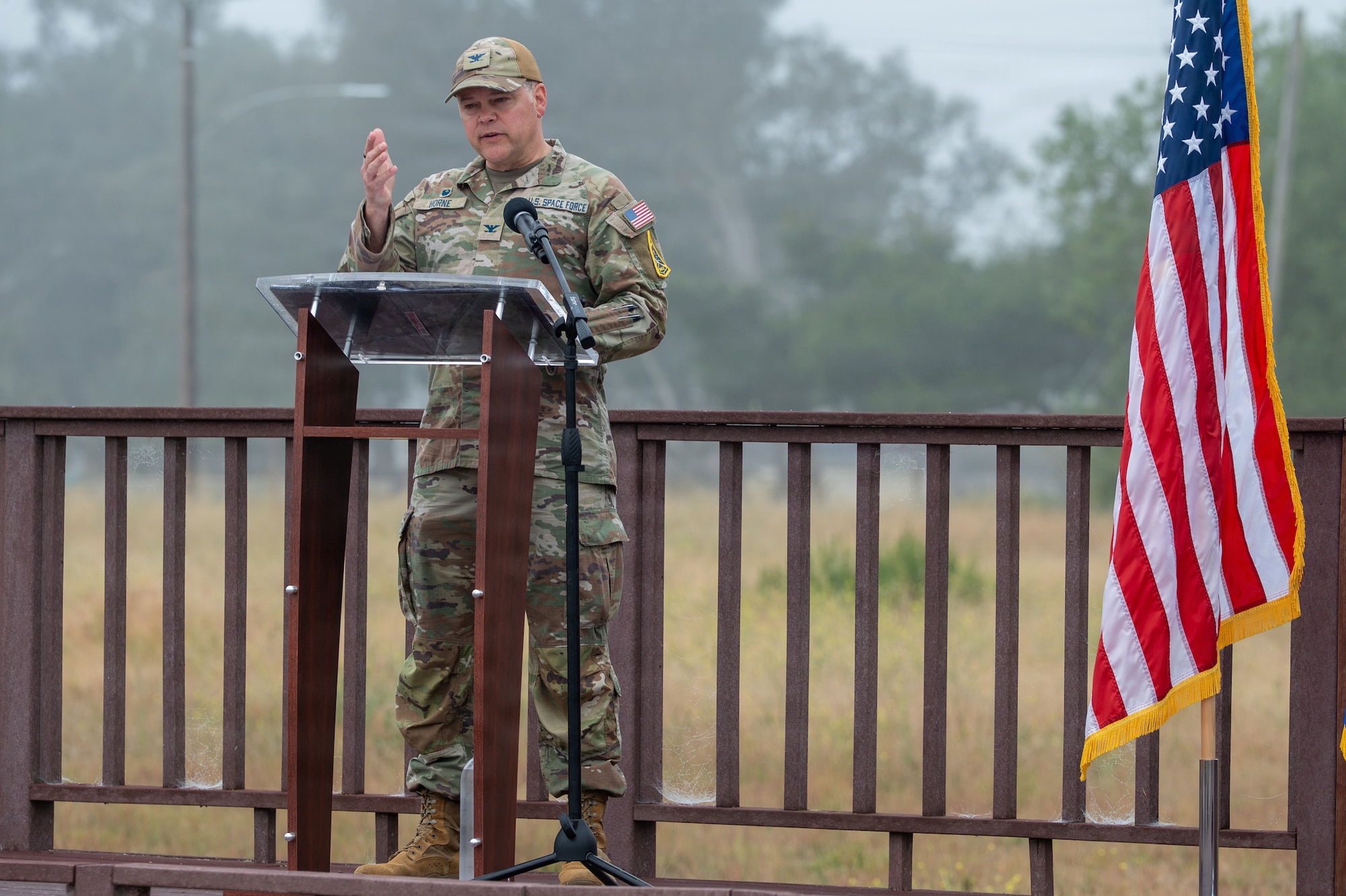 U.S. Space Force Col. James T. Horne III, Space Launch Delta 30 commander, motions to the crowd as he stands and speaks behind a podium on stage outside.