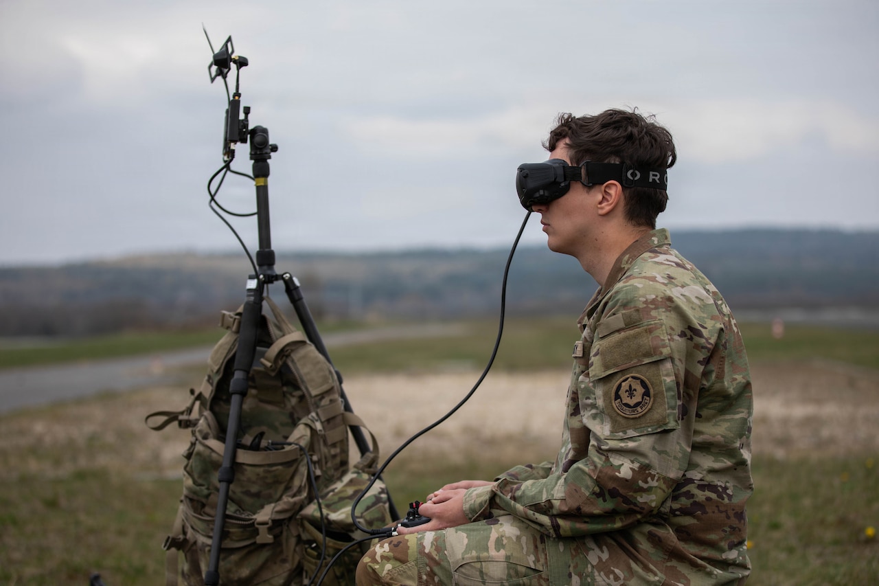 A man in a camouflage military uniform wears goggles attached to a remote control outside in a grassy field; next to him is a tripod with a camouflage backpack hanging on it.