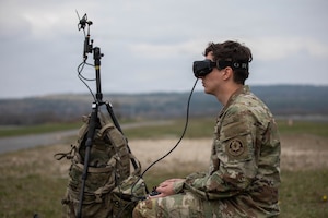 A man in a camouflage military uniform wears goggles attached to a remote control outside in a grassy field; next to him is a tripod with a camouflage backpack hanging on it.