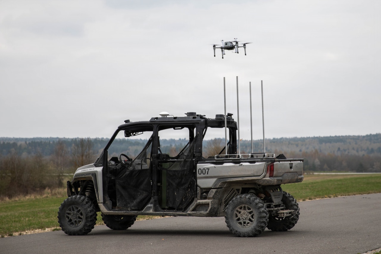 A drone takes off from an unmanned ground vehicle parked on a road near a grassy field with trees in the distance.