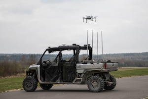 A drone takes off from an unmanned ground vehicle parked on a road near a grassy field with trees in the distance.