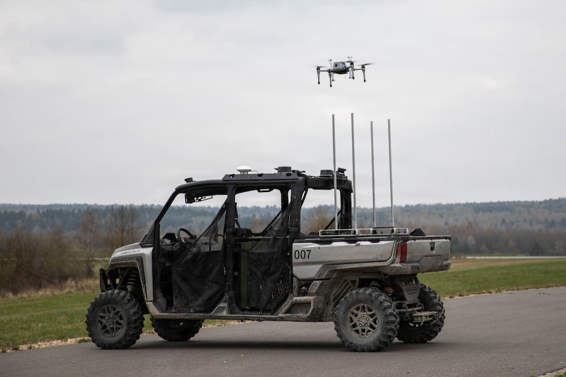A drone takes off from an unmanned ground vehicle parked on a road near a grassy field with trees in the distance.