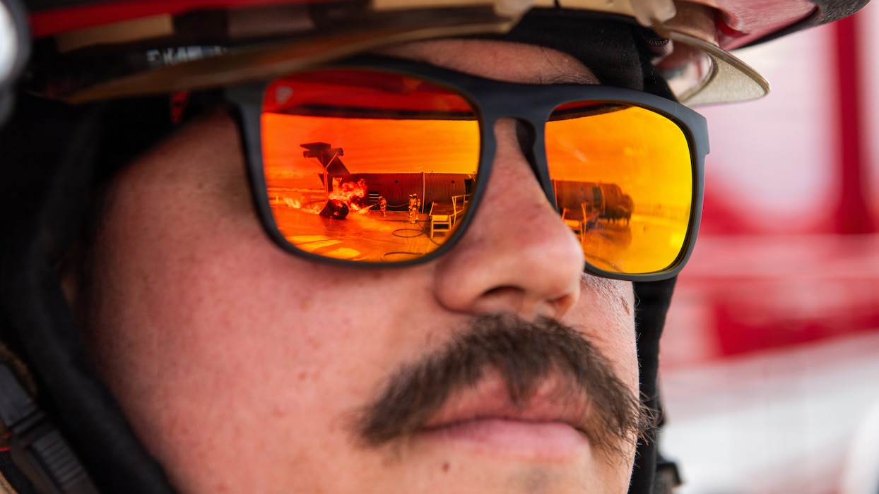 A firefighter's glasses reflect firefighters combat a simulated aircraft fire