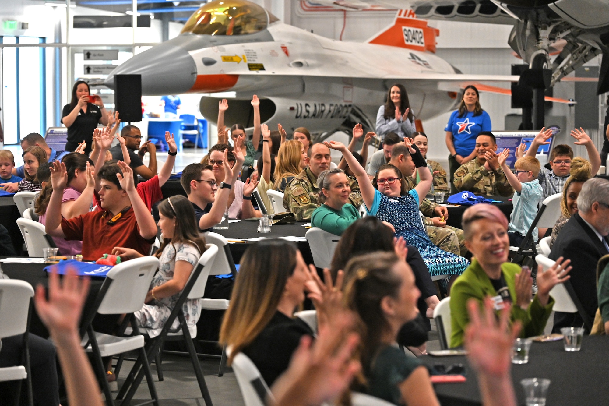 A diverse audience of military families, including several young children raising their hands, are seated inside the Hill Aerospace Museum gallery.