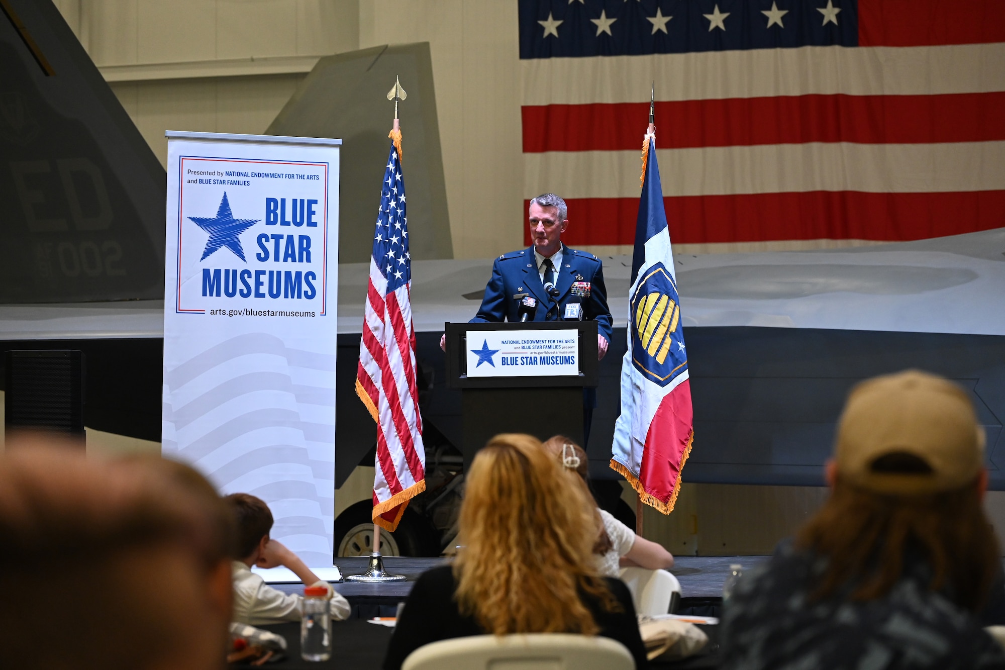Col. Daniel Cornelius, in a U.S. Air Force uniform, stands at a lectern speaking into a microphone.