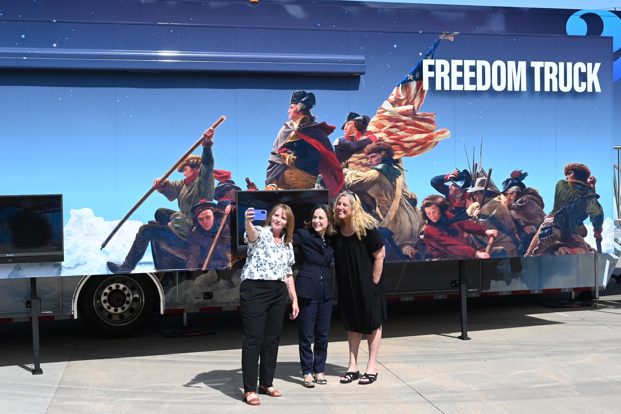 Three event organizers smile while posing for a group selfie in front of the Freedom 250 Mobile Museum trailer.