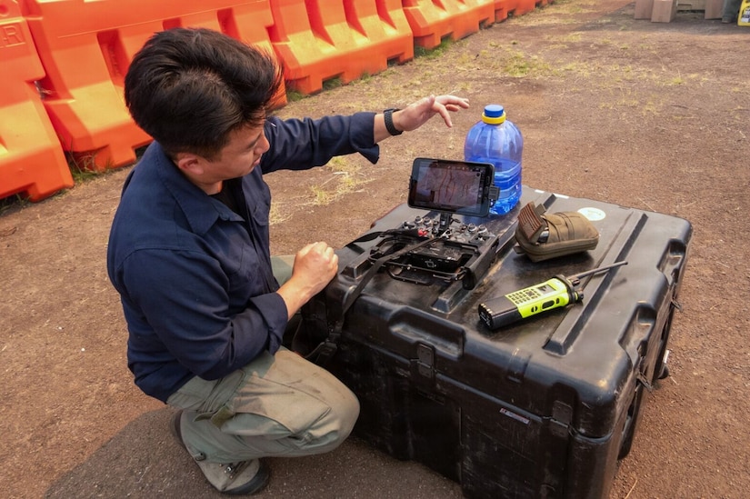 A man in casual attire kneels while showing a drone remote sitting on a container outside.