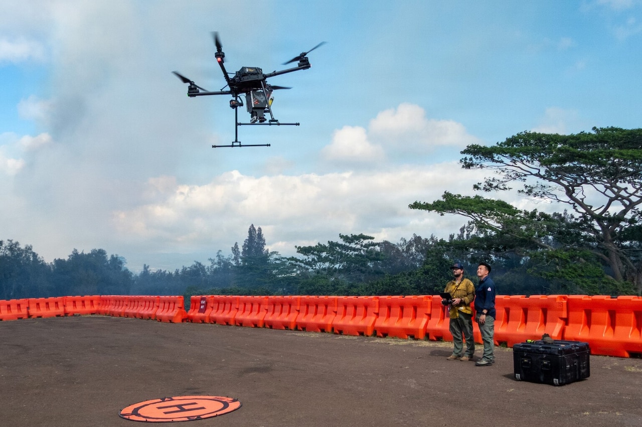 A man in casual attire uses a remote to fly a drone, while another man in similar attire stands next to him outside in an open area; there are trees in the background and a protective barrier lining the area.