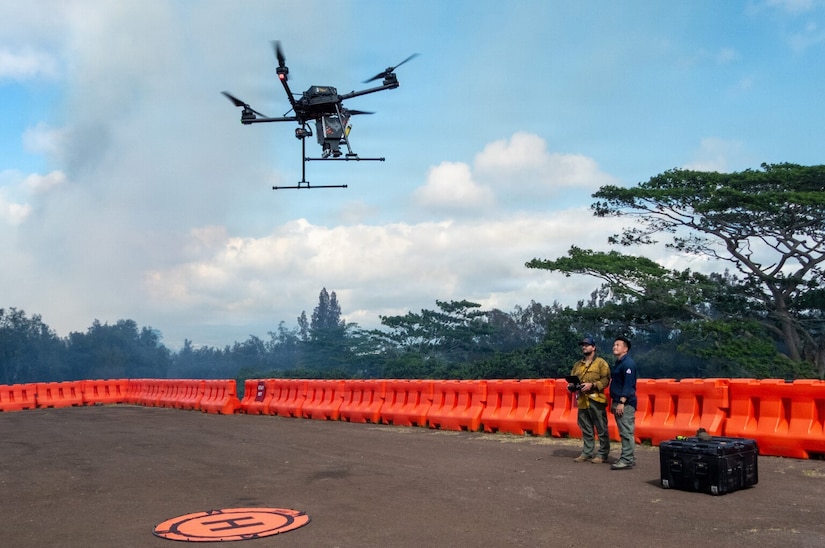 A man in casual attire uses a remote to fly a drone, while another man in similar attire stands next to him outside in an open area; there are trees in the background and a protective barrier lining the area.
