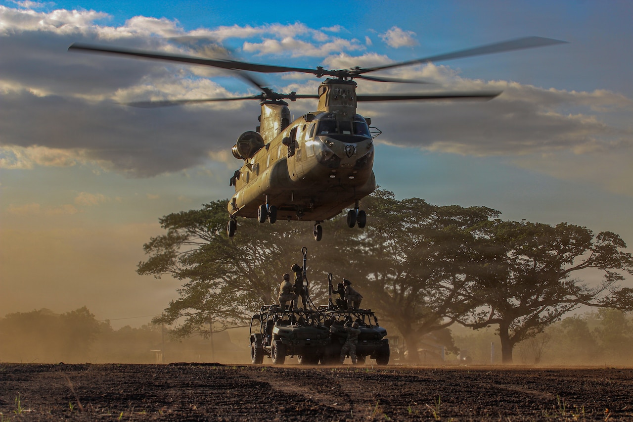 Four service members wearing camouflage military uniforms work to attach hooks tethered to two military vehicles to the bottom of a military helicopter that hovers above them.