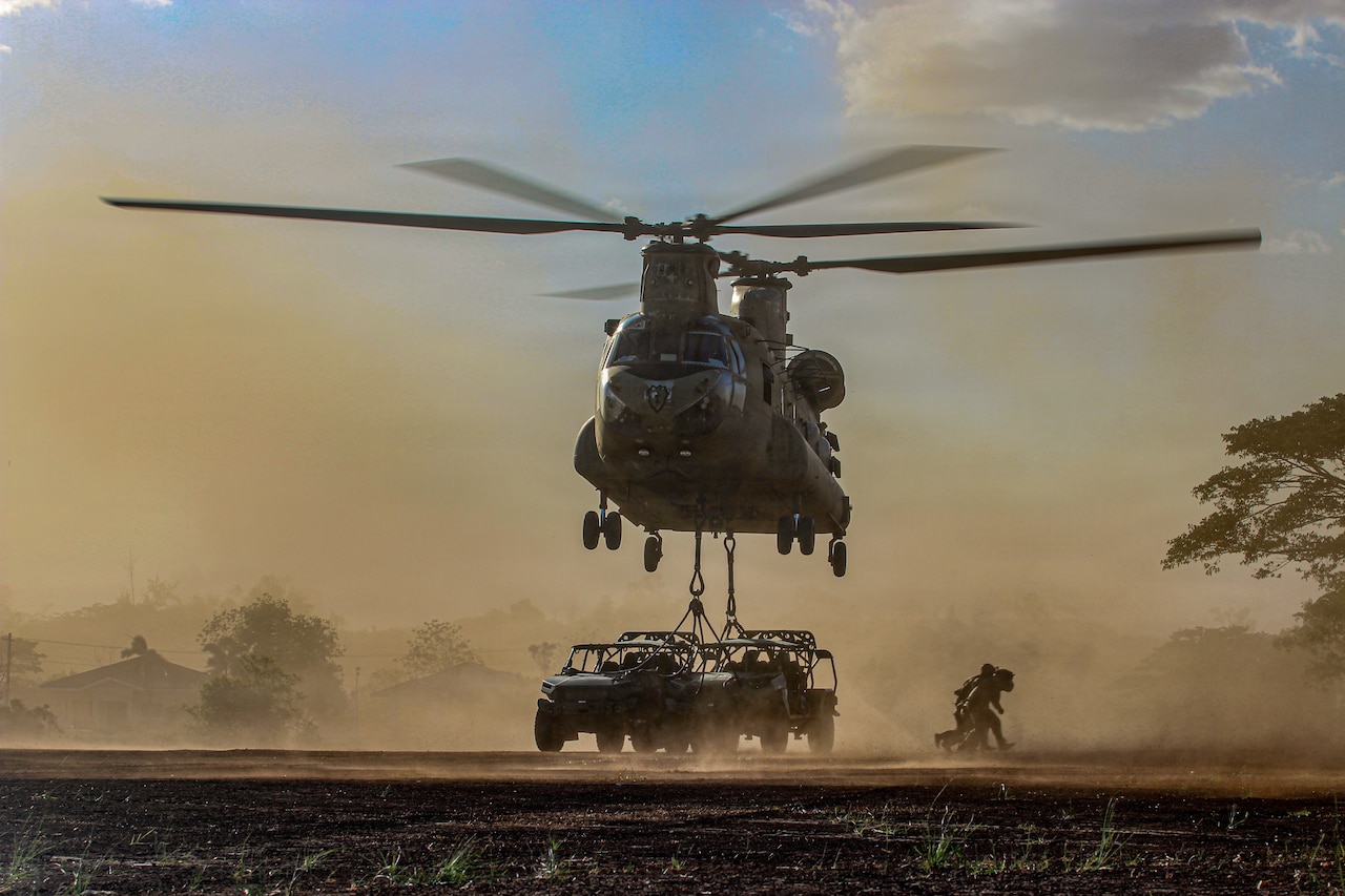 A group of service members wearing camouflage military uniforms run away from a military helicopter that is attached to two military vehicles as red dust surrounds them.