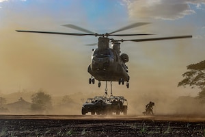 A group of service members wearing camouflage military uniforms run away from a military helicopter that is attached to two military vehicles as red dust surrounds them.