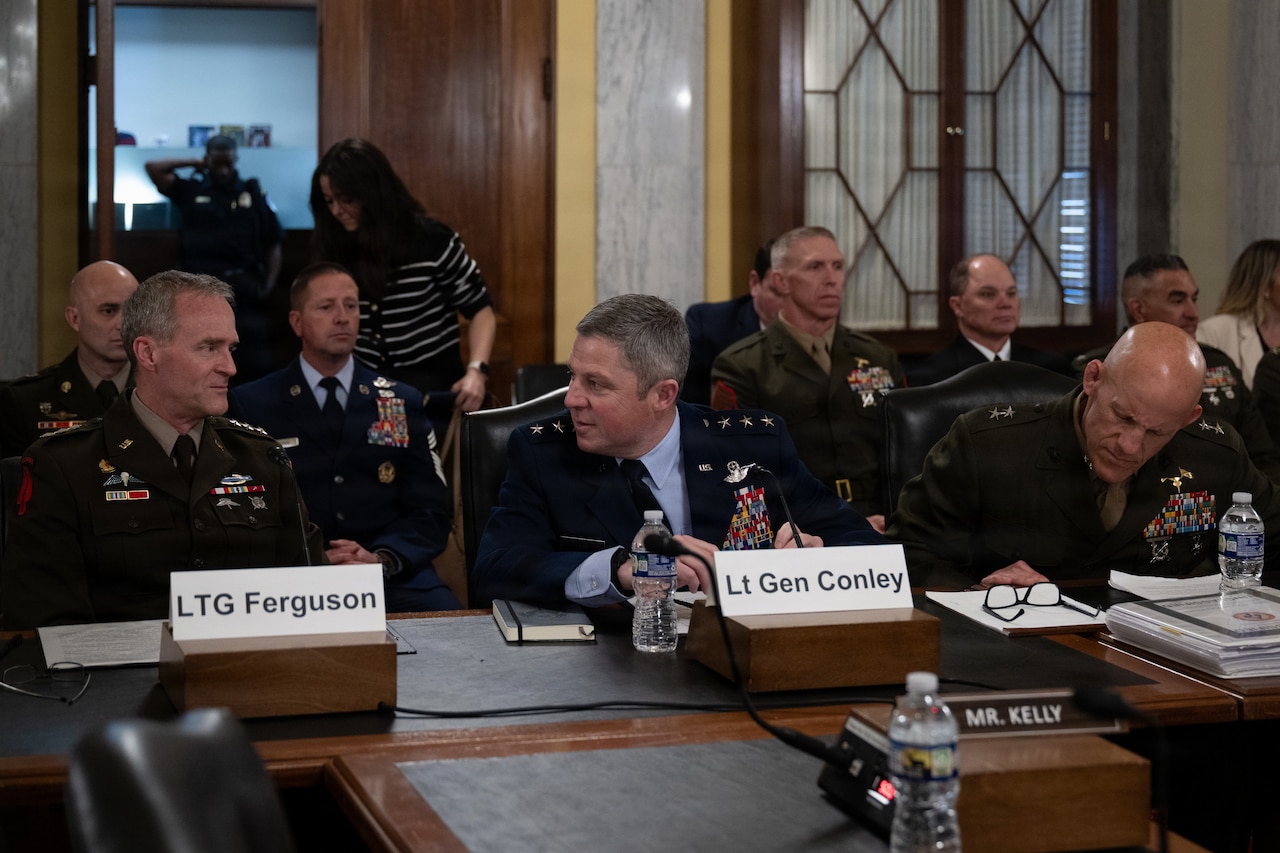 Three men wearing dress military uniforms sit indoors at a long table with microphones in front of them. Sitting behind them are two rows of other people, some of them in uniform and some in civilian business attire.