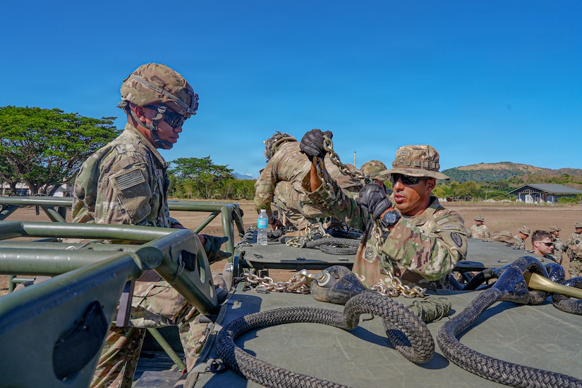 A service member wearing a camouflage military uniform holds a large chain as another service member in similar attire stands on a military vehicle and looks on.
