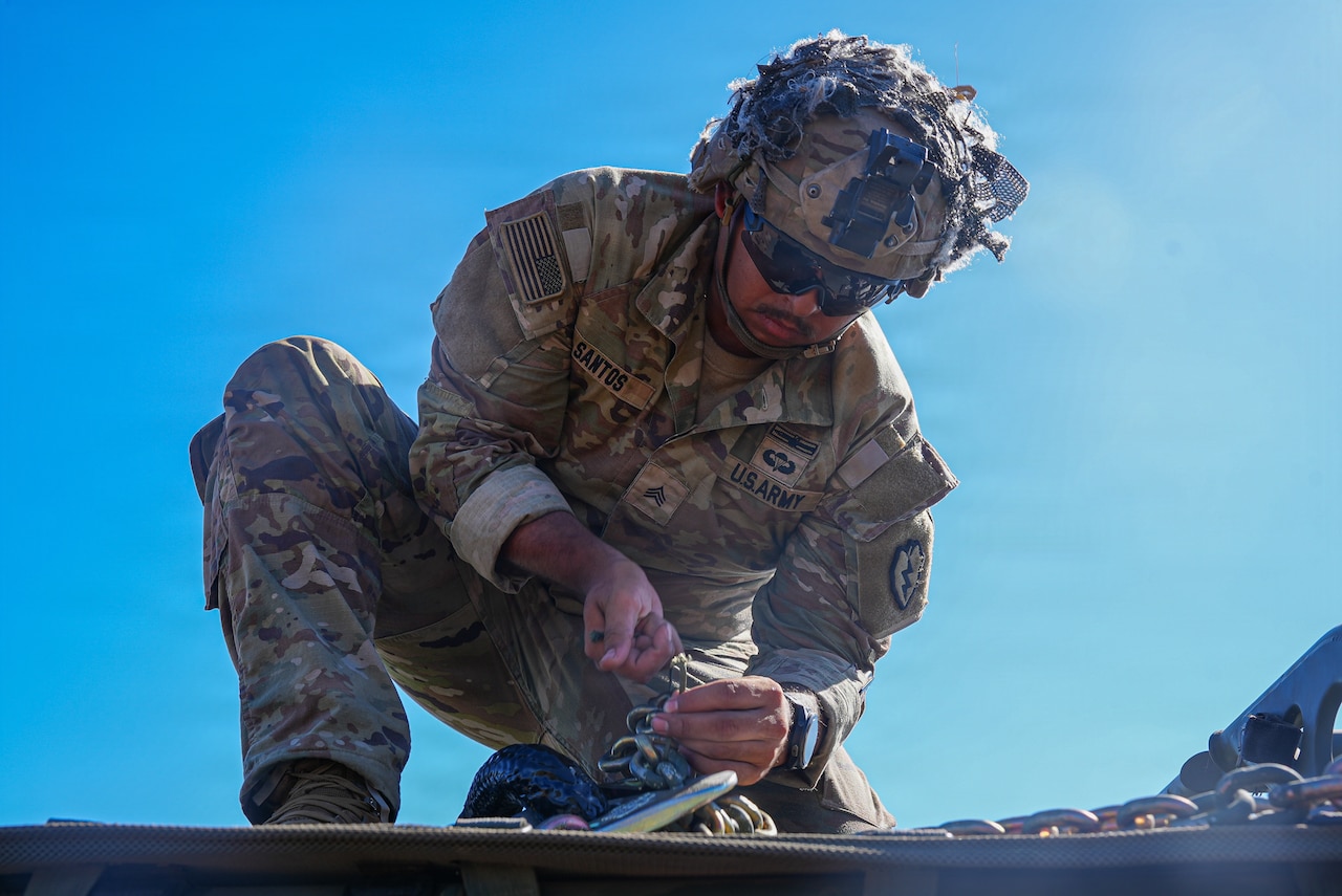 A service member wearing a camouflage military uniform connects a metal hook to a military vehicle.