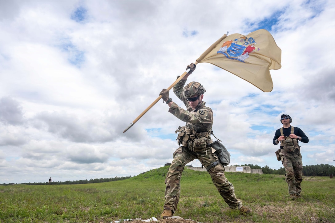 A soldier prepares to put a flag into a field during the day as a fellow soldier watches from behind.