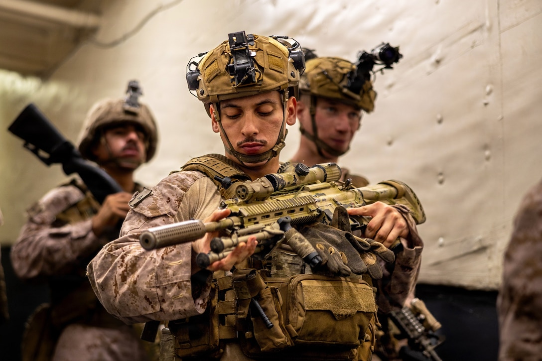 A Marine in tactical gear looks at their weapon while fellow Marines walk nearby.