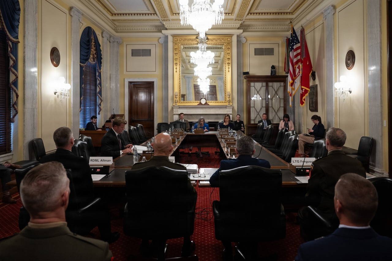 A group of people are sitting around a large, wooden table in an ornate room. Glass chandeliers hang from the ceiling.