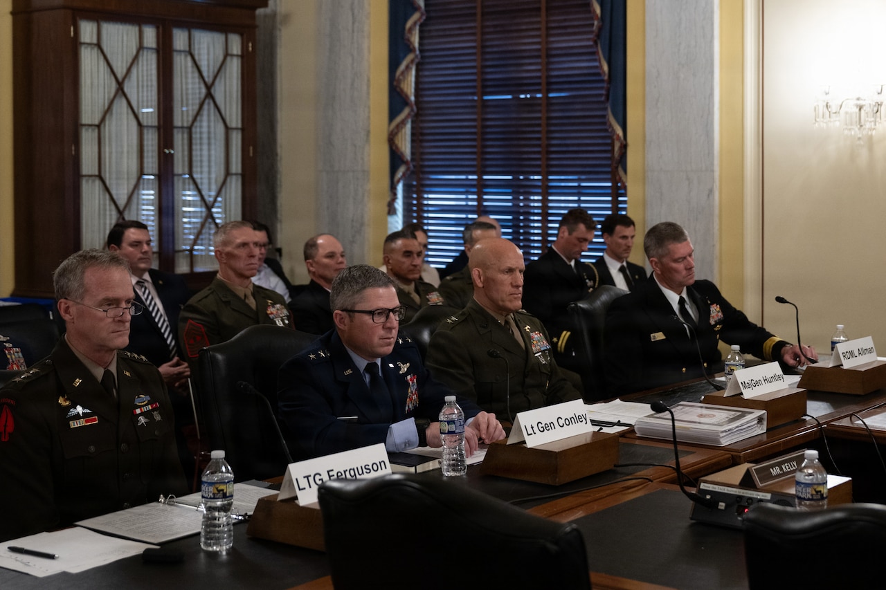 Four men wearing dress military uniforms sit indoors at a long table with microphones in front of them. Sitting behind them are two rows of other people, some of them in military uniforms and some in civilian business attire.