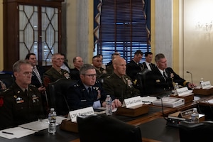 Four men wearing dress military uniforms sit indoors at a long table with microphones in front of them. Sitting behind them are two rows of other people, some of them in military uniforms and some in civilian business attire.