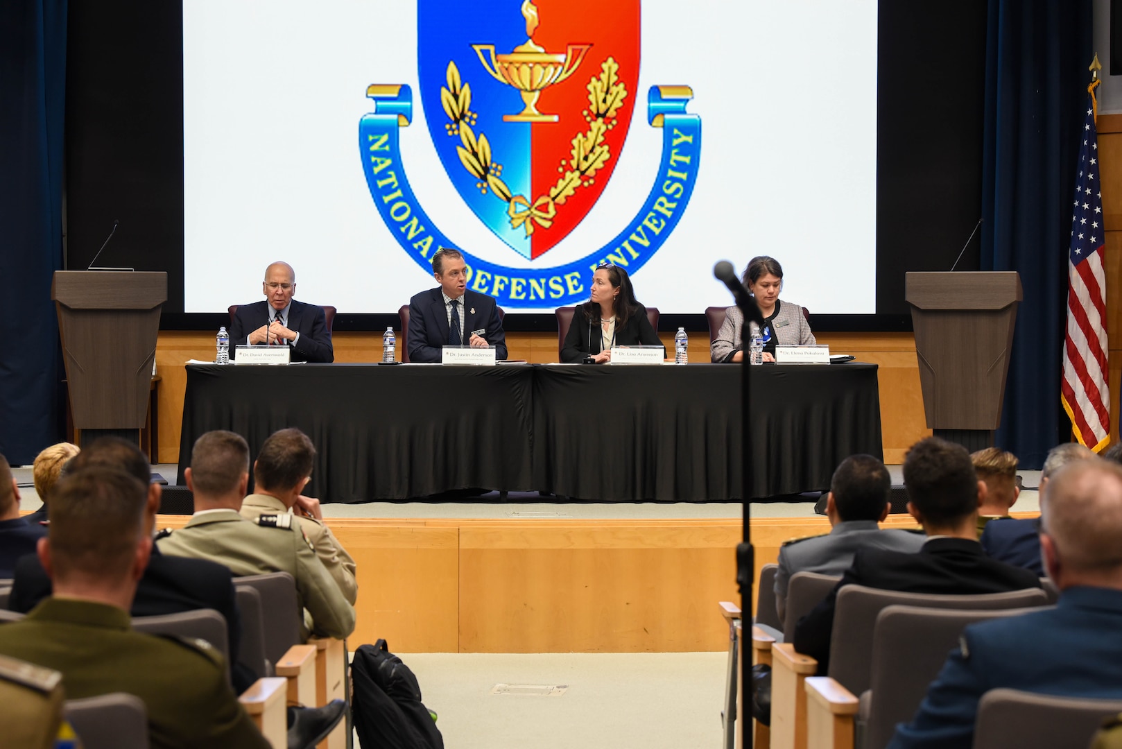 Speakers part of the "U.S. National Defense Strategy and its Implications for NATO and Europe" panel sit in front of a large screen with NDU's logo. From left to right: Dr. David Auerswald, Professor, National War College, NDU; Dr. Justin Anderson, Senior Fellow, INSS, NDU; Dr. Lisa A. Aronsson, Research Fellow, Institute for National Strategic Studies (INSS), NDU (Moderator); and Dr. Elena Pokalova, Professor, College of International Security Affairs (CISA).