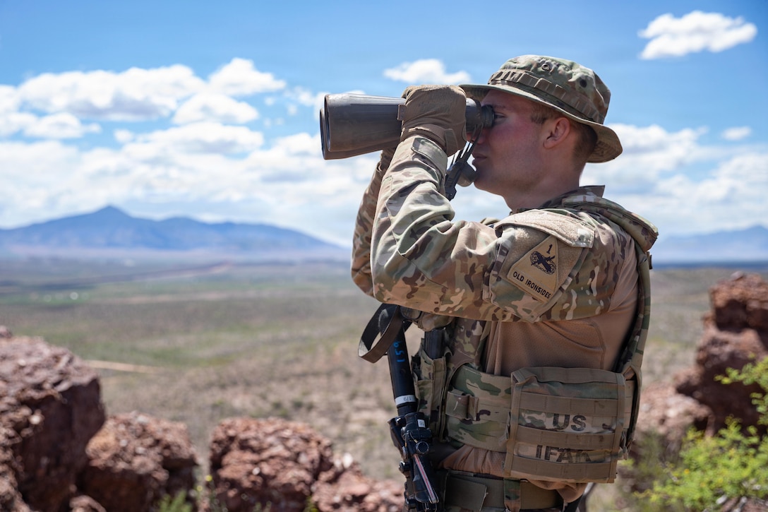 A soldier stands and looks west through binoculars, with large red rocks, blue sky and mountains in the background.