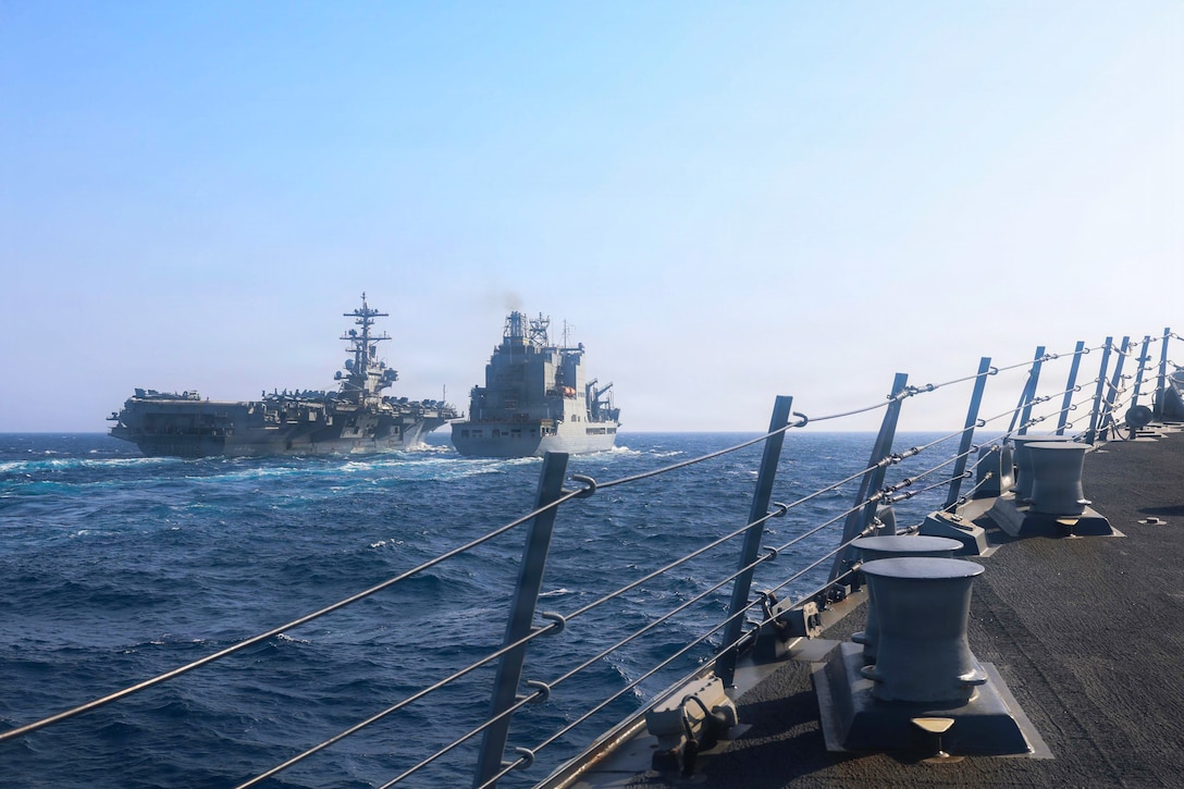 Two military ships are seen from the deck of a third, with the horizon in the background.