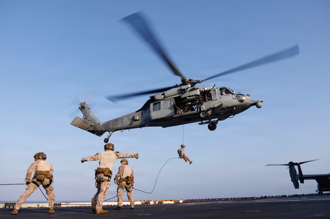 A Marine hangs from a rope out of a helicopter hovering above a ship's deck, where two Marines hold the rope and a third makes arm motions.