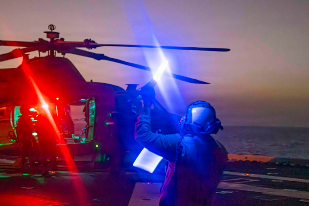 A helicopter is parked on the deck of a ship at night, with red light reflected on two people standing near its doorway and blue light reflected on a person signaling to them.
