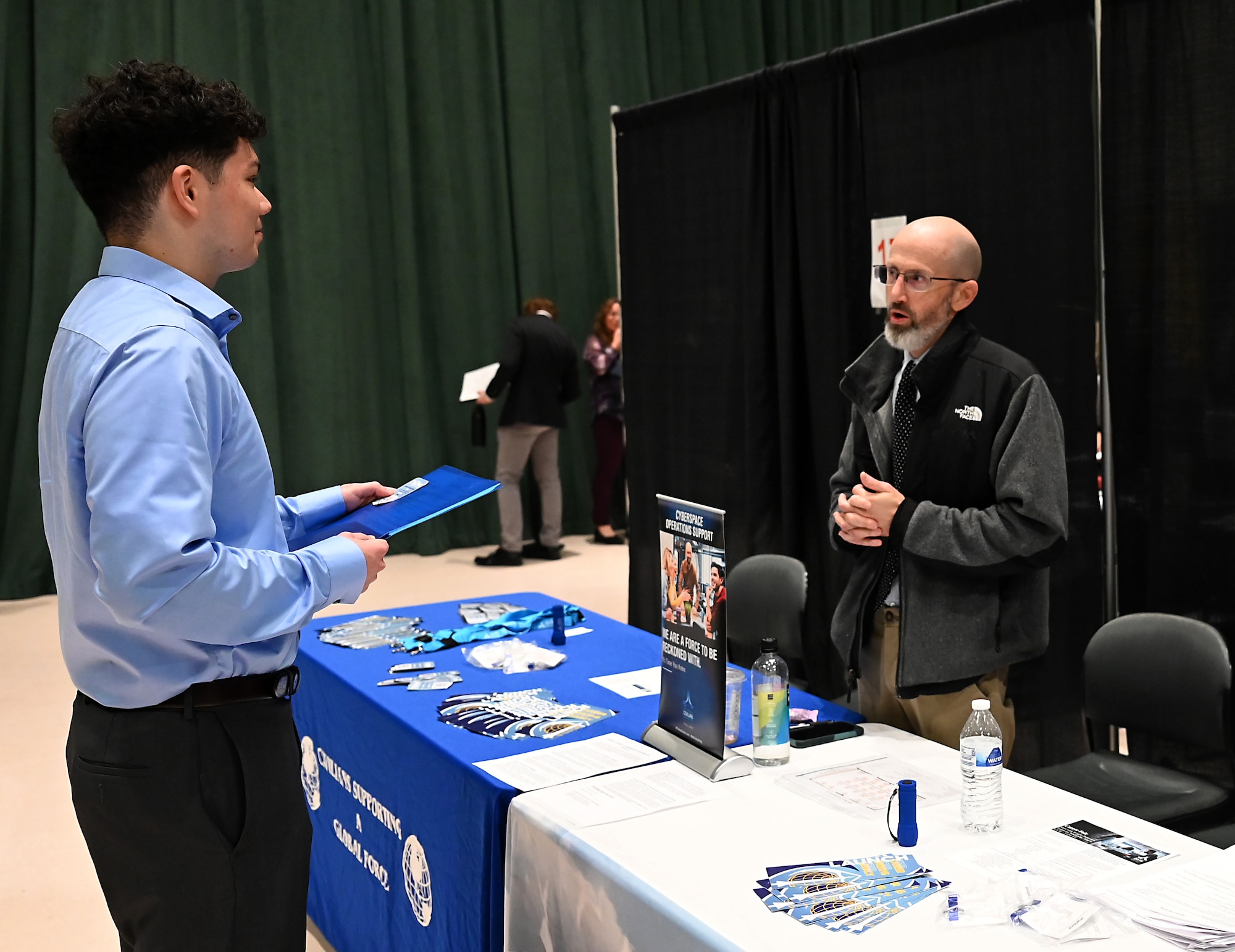 A bald man describes something to a younger male event attendee in a button up shirt.