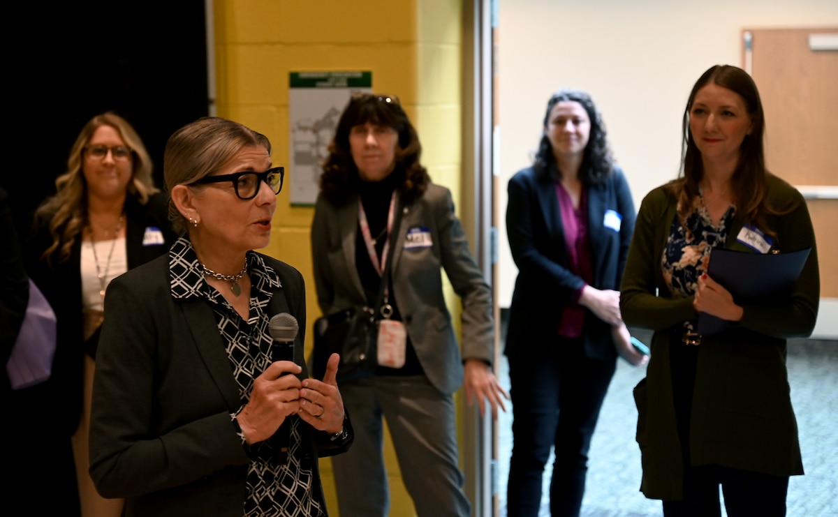 A woman in a business suit addresses a group of four other women in business suits.