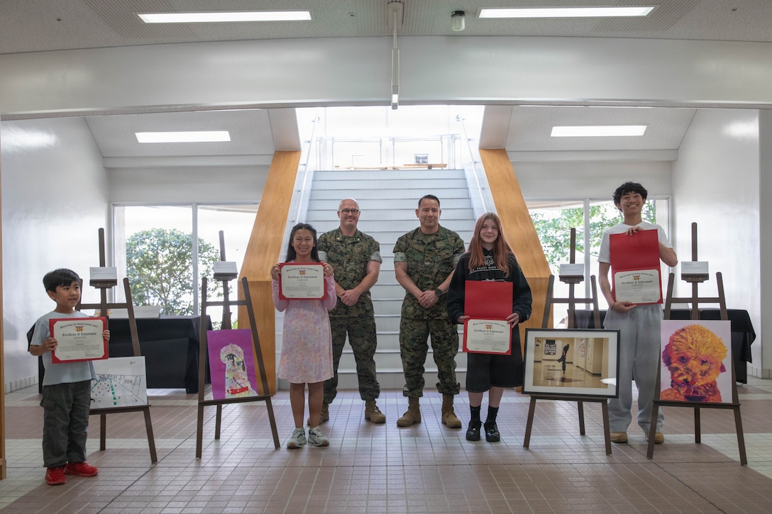 U.S. Marine Corps Maj. Mason Englehart, middle left, the executive officer of Headquarters and Headquarters Squadron (HHS), and a native of Murfreesboro, Tennessee, and U.S. Marine Corps Sgt. Maj. Richard Johnson, the sergeant major of Marine Corps Air Station Iwakuni, and a native of Washington D.C., pose for a group photo with the winners of the May art awards during the art awards presentation at MCAS Iwakuni, Japan, May 11, 2026. The air station presented certificates for its monthly art competition in which students attending schools at the air station showcase their artistic talent through different mediums. (U.S. Marine Corps photo by Lance Cpl. Gage Collins)