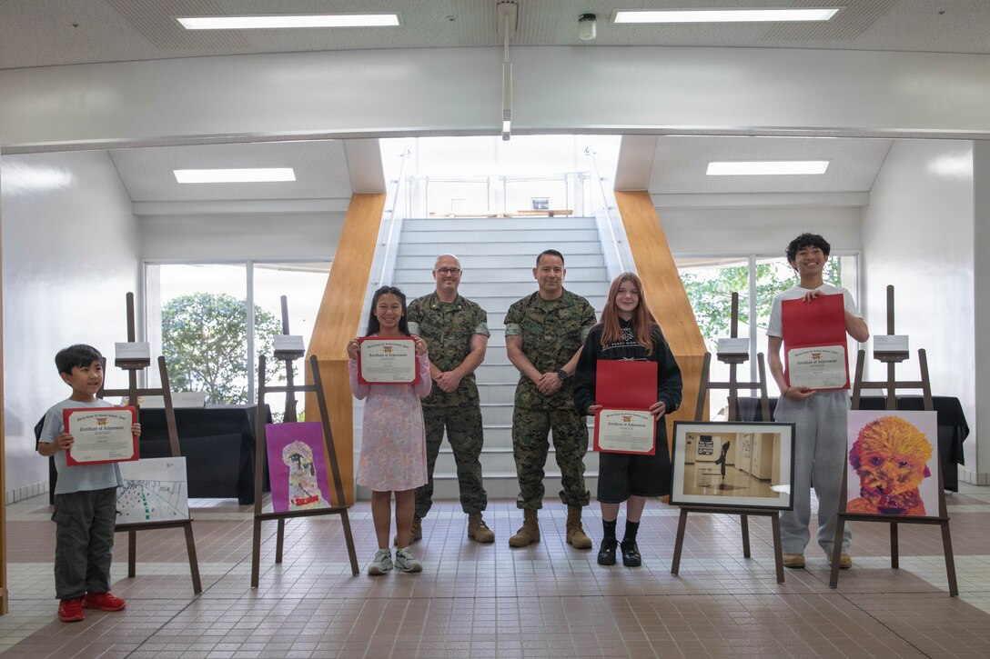 U.S. Marine Corps Maj. Mason Englehart, middle left, the executive officer of Headquarters and Headquarters Squadron (HHS), and a native of Murfreesboro, Tennessee, and U.S. Marine Corps Sgt. Maj. Richard Johnson, the sergeant major of Marine Corps Air Station Iwakuni, and a native of Washington D.C., pose for a group photo with the winners of the May art awards during the art awards presentation at MCAS Iwakuni, Japan, May 11, 2026. The air station presented certificates for its monthly art competition in which students attending schools at the air station showcase their artistic talent through different mediums. (U.S. Marine Corps photo by Lance Cpl. Gage Collins)