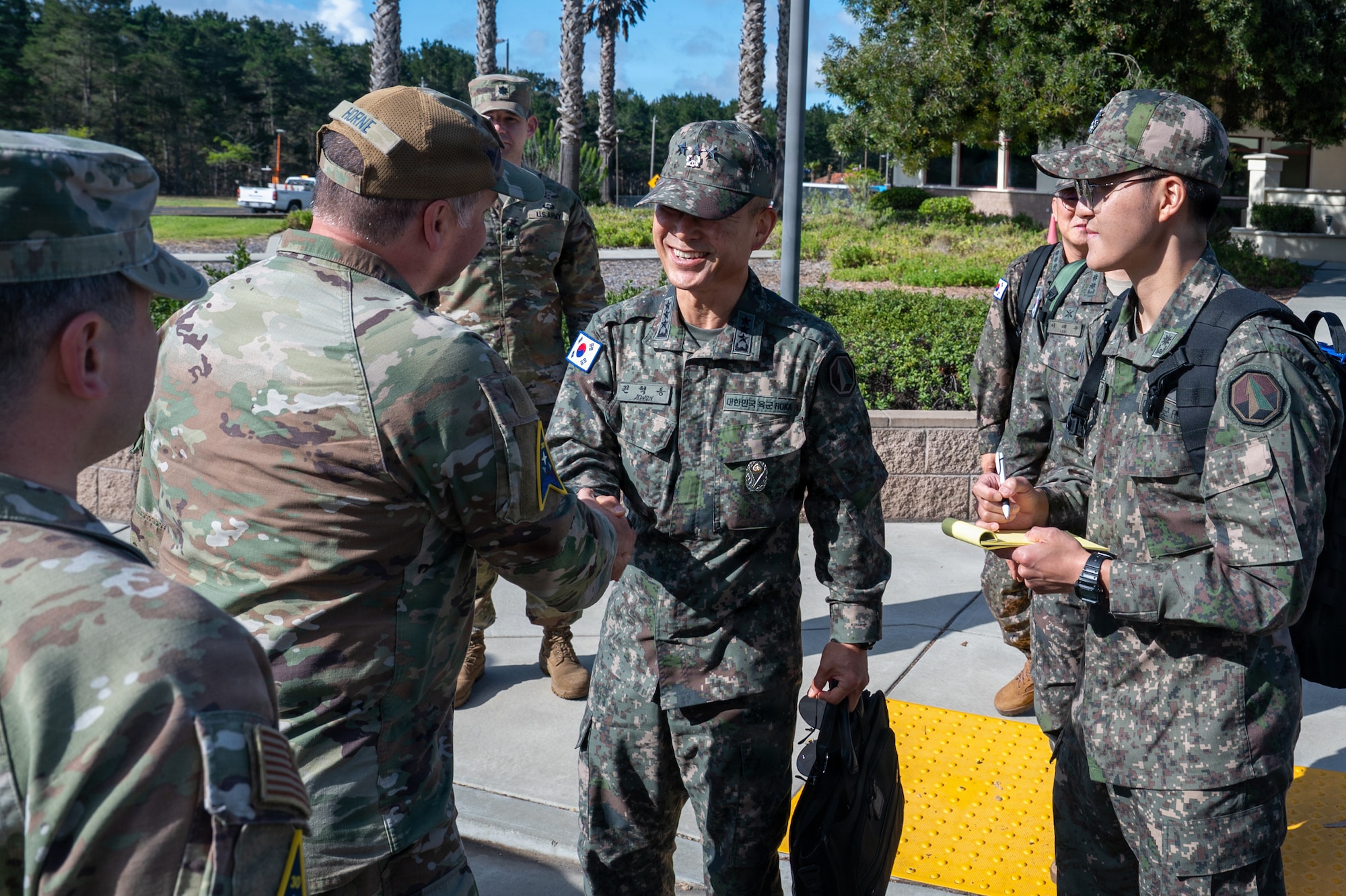 Two people shake hands while others watch outside a gate entrance building.