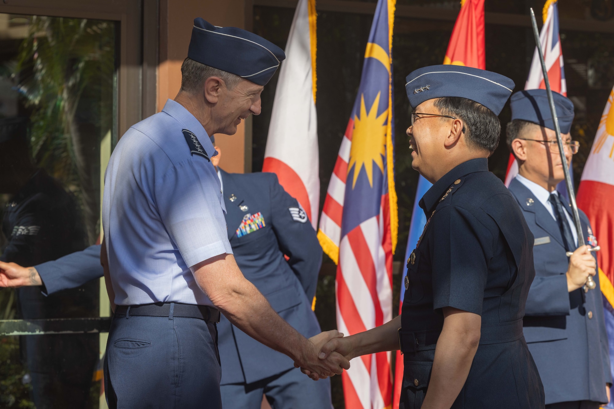U.S. Pacific Air Forces commander, Gen. Kevin Schneider, and Philippine Air Force commanding general, Lt. Gen. Arthur Cordura, smile as they shake hands. They are standing in front of a vibrant backdrop of international flags, including those of Japan, Malaysia, and the Philippines.
