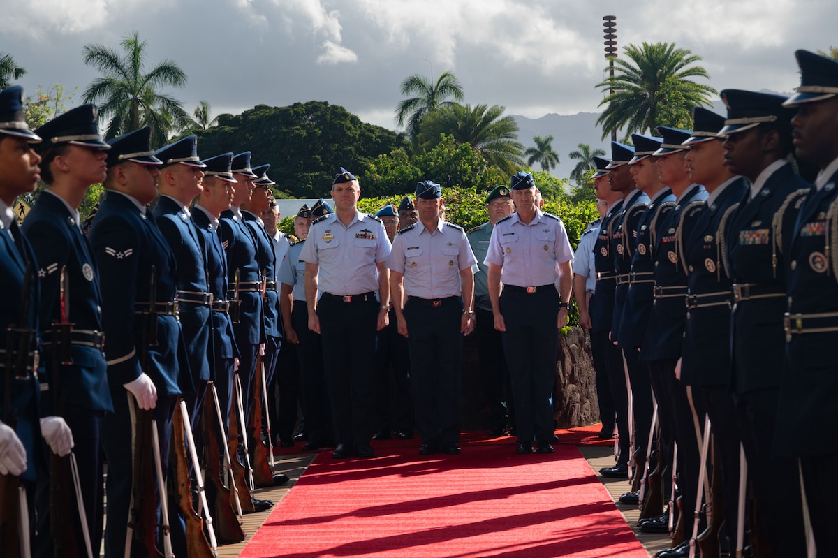 U.S. Air Force Chief of Staff Gen. Ken Wilsbach, U.S. Air Force Gen. Kevin Schneider, Pacific Air Forces commander, and Royal Australian Air Force Air Marshal Stephen Chappell, Chief of Air Force, stand together in uniform at the end of a red carpet. They are framed by two rows of an honor cordon made up of Airmen in dress uniforms, with a tropical background of palm trees and mountains.